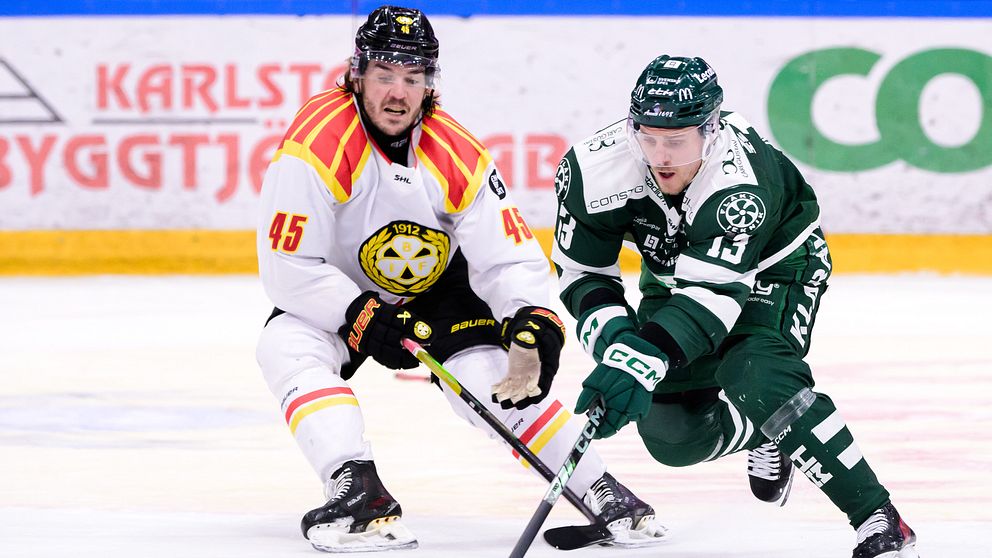 Två ishockeyspelare i matchdräkter från olika lag kämpar om pucken på isen, en i vita och röda färger med nummer 45 och den andra i gröna med nummer 15. Bakgrunden visar en del av ishockeyrinken med reklam.