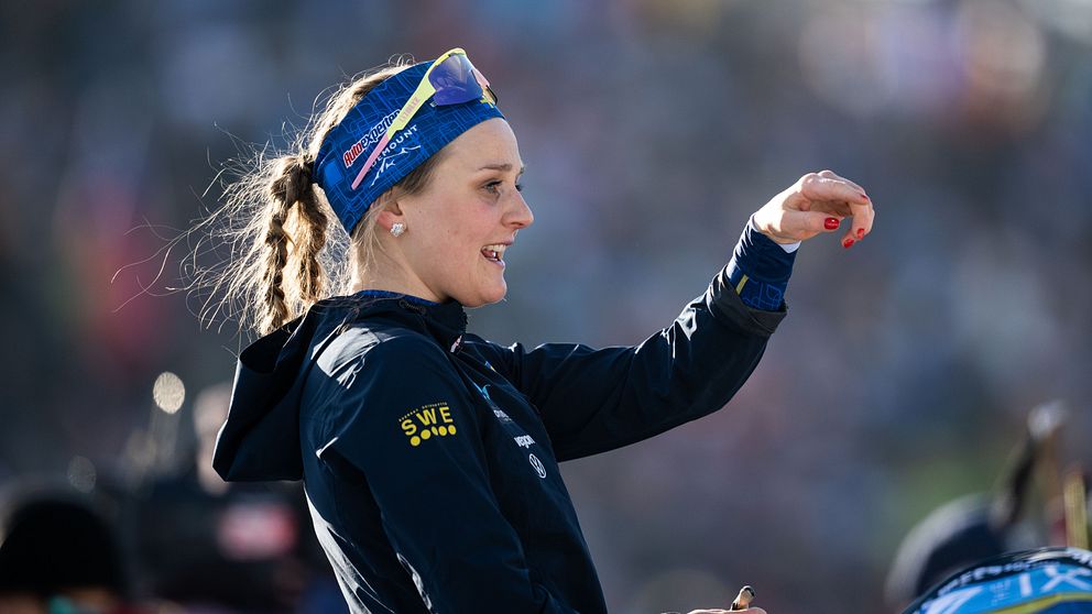 En person i en sportjacka som visar en gest av koncentration eller anvisning, bärande en hjälm och goggles, står på en tävling med en stor publik i bakgrunden.