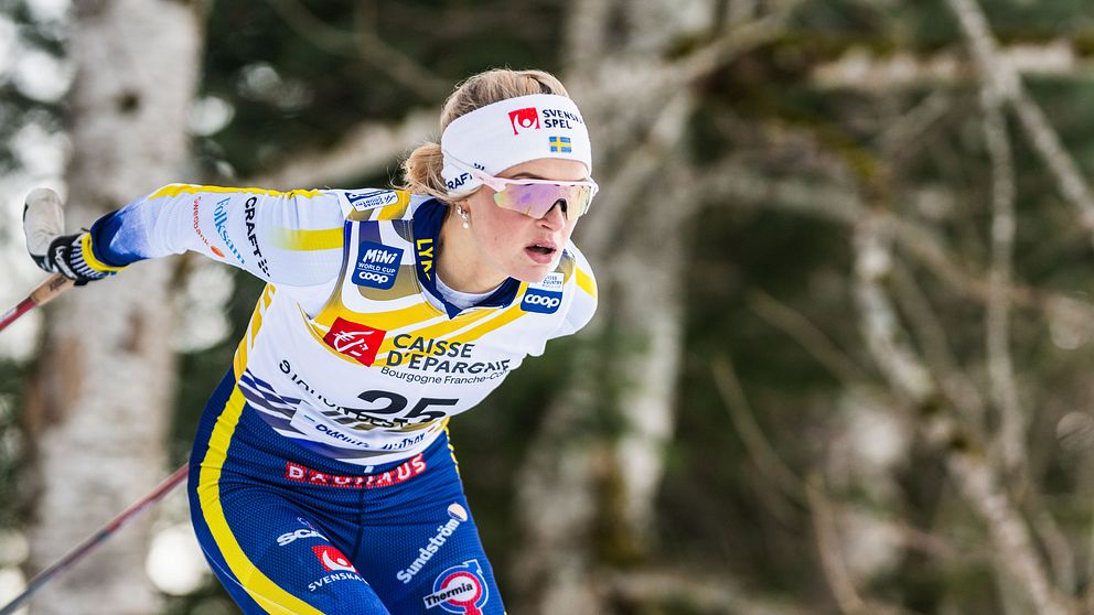 En skidåkare i tävlingsdräkt ställer sig i en dynamisk position under en vintersporttävling, med träd i bakgrunden som skapar en naturskön miljö.