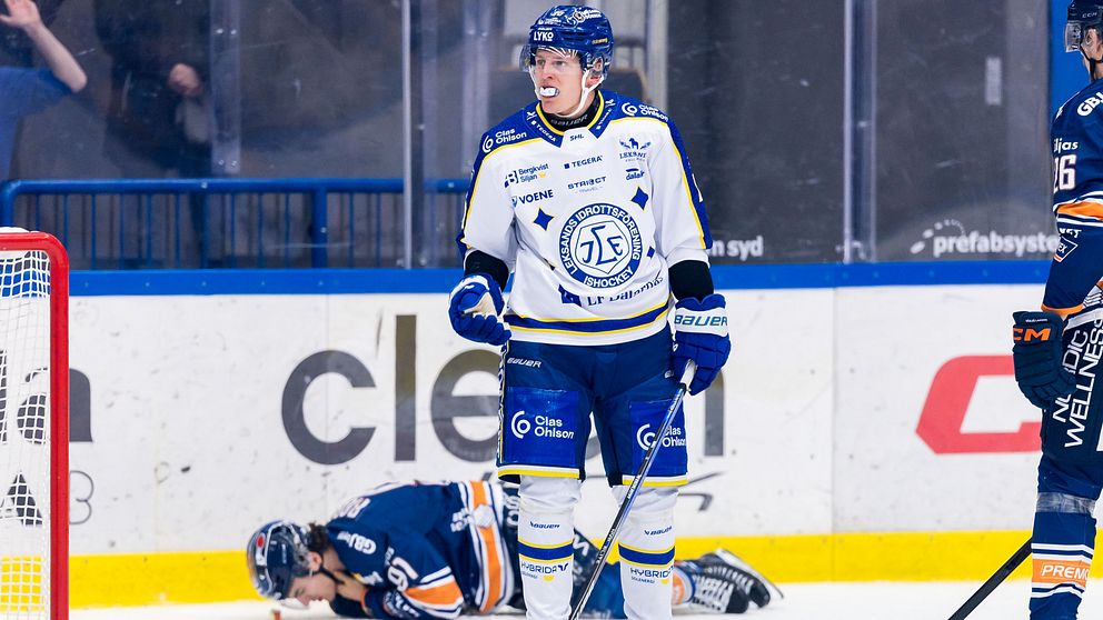 En ishockeyspelare i blått och vitt står i förgrunden medan en annan spelare ligger på isen i bakgrunden efter en kollision. Det är en match på en inomhusarena med tydlig fokus på spelarnas reaktioner.