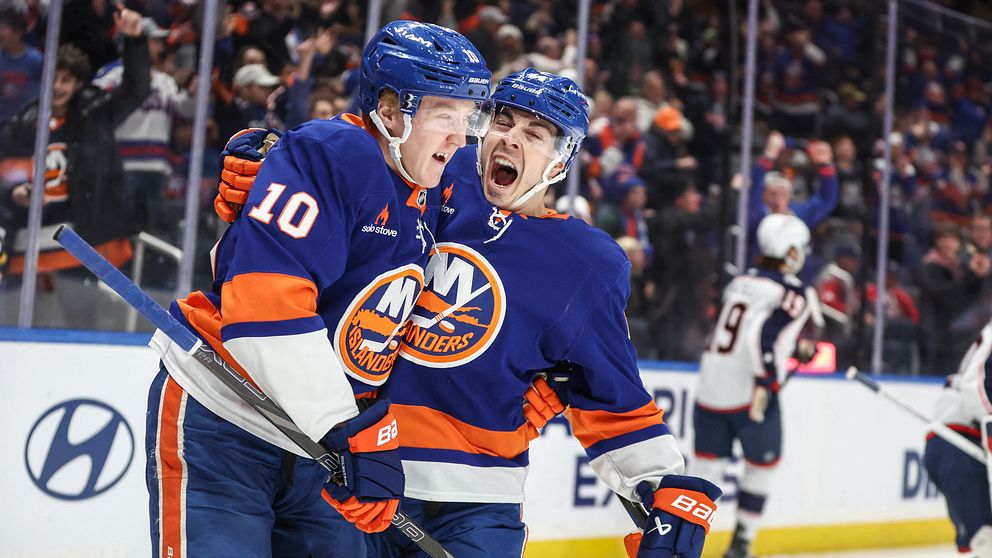Två hockeyspelare från New York Islanders omfamnar varandra i firande på isen med en motspelare från motståndarlaget skymtande i bakgrunden. Publiken syns på läktarna.