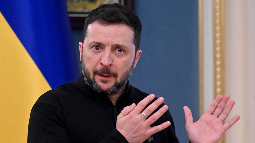 Person gestikulerar framför ukrainska flaggan i ett formellt rum.