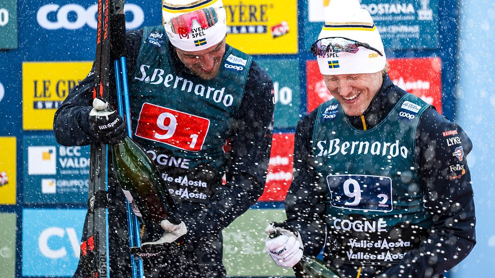Två personer i skidkläder står framför en prisscen dekorerad med sponsormärken. De håller skidor och en champagneflaska, och snö faller omkring dem.