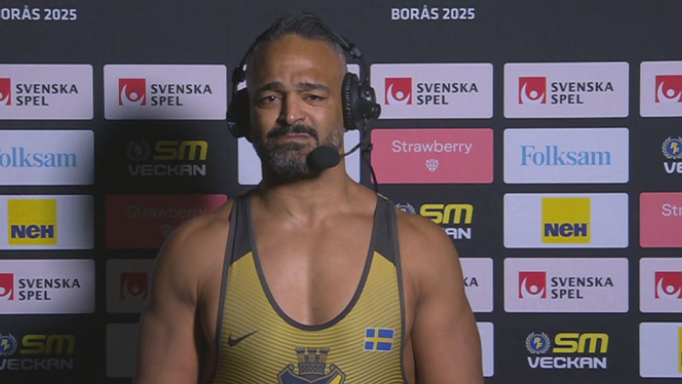 En brottare i AIK:s tävlingsdräkt står framför en bakgrund med olika logotyper och sponsormärken vid ett evenemang.