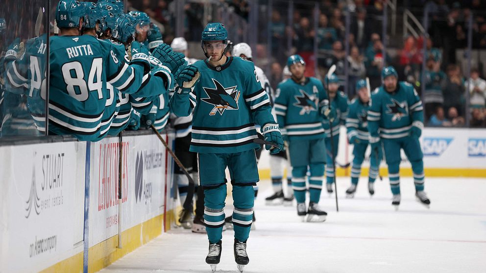 Spelare från San Jose Sharks i sina turkosa matchtröjor firar ett mål vid bänken under en ishockeymatch. I bakgrunden syns fler lagmedlemmar och en fullsatt arena. Atmosfären är energisk och fansen är engagerade.