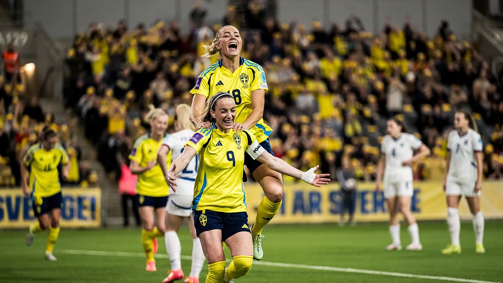 En svensk fotbollsspelare firar ett mål med glädje, där hon står på ryggen av en medspelare. I bakgrunden syns en stor publik iförd gula kläder, vilket skapar en energisk atmosfär.