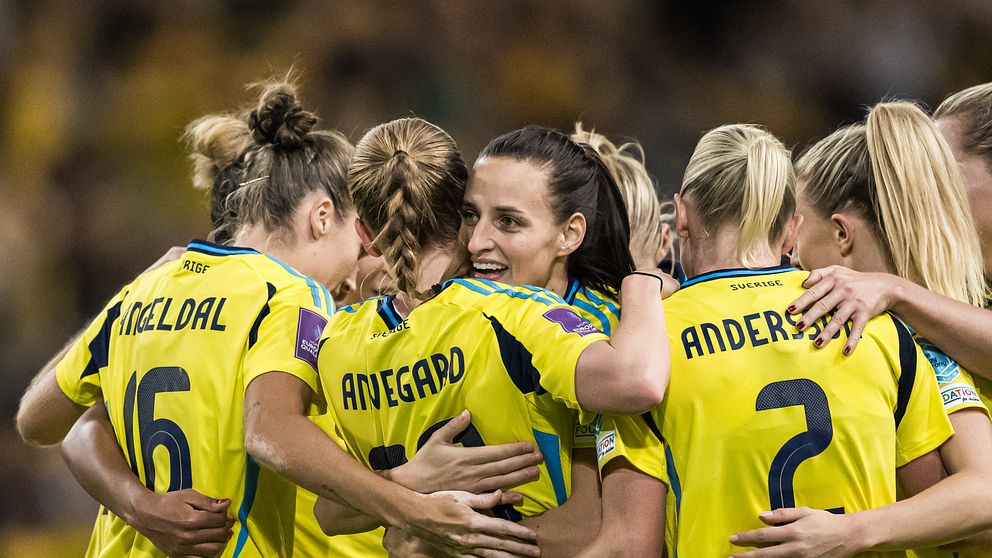 Fem kvinnliga fotbollsspelare står tillsammans i en stöttande omfamning, iförda gula tröjor med blå detaljer. De är samlade efter ett mål eller en framgång, med fokus på laganda och kamratskap. Bakgrunden visar suddiga åskådare som skapar en energifylld atmosfär.