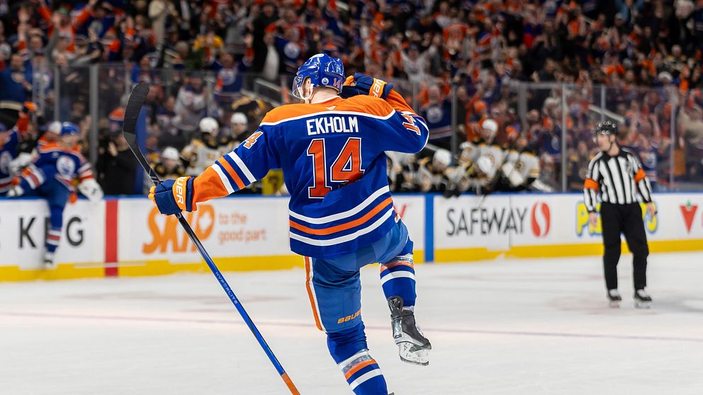 Spelaren Mattias Ekholm från Edmonton Oilers firar efter att ha gjort mål, med en energisk gest där han sätter handen på huvudet. I bakgrunden jublar publiken medan en domare observerar spelet.