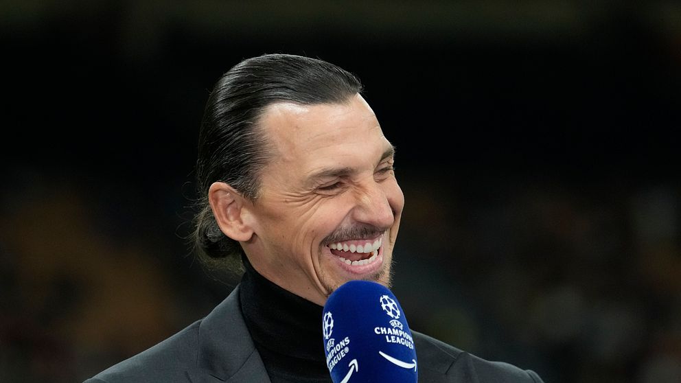 En person håller i en mikrofon med Champions League-loggan på, iklädd en mörk kavaj och svart polotröja. Bakgrunden är oskarp med dunkla färger, vilket antyder att bilden är tagen i en inomhusmiljö, möjligen vid ett sportevenemang.