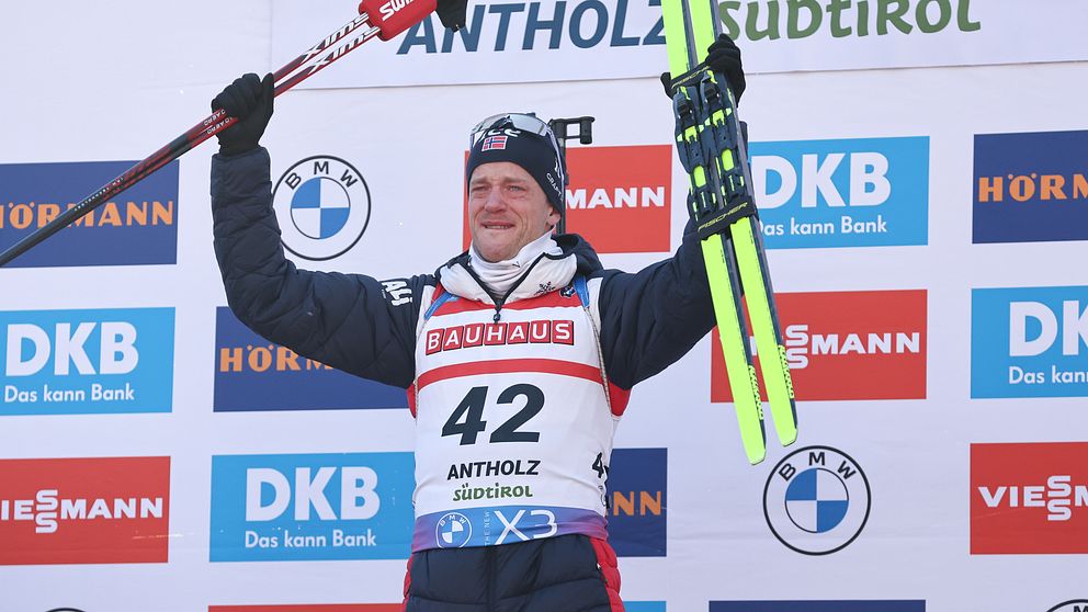 En skidåkare klädd i en tävlingsdräkt och nummerlapp 42, firar med skidor i ena handen och en skidstav i den andra framför en bakgrund fylld med sponsormärken som ”ANTHOLZ”, ”BMW”, ”DKB” och ”HÖRMANN”.