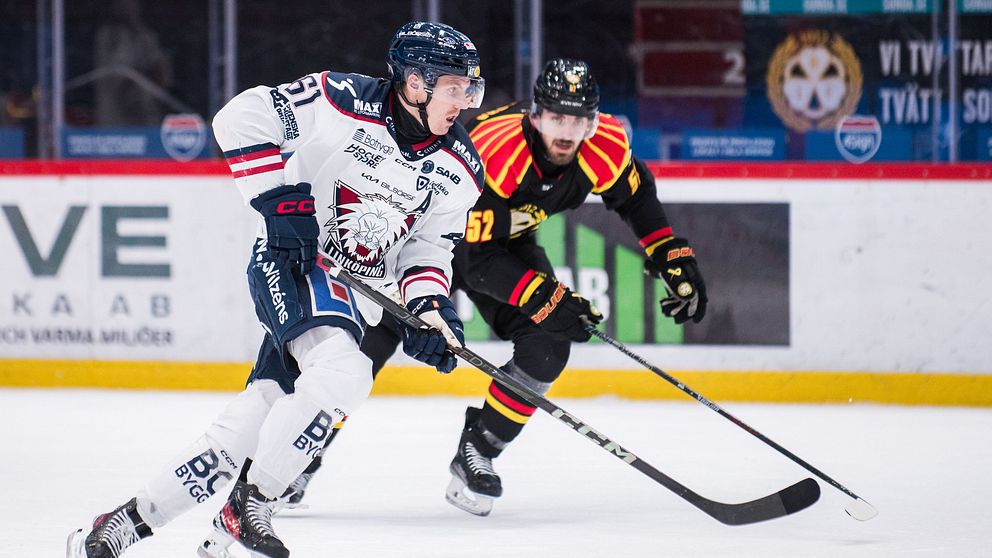 Två ishockeyspelare från motståndarlag jagar pucken under en match. Spelaren i vita kläder åker snett framför den andra spelaren som har svarta och röd-guldfärgade dräkter.