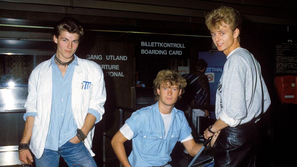 Tre unga vuxna i 1980-talskläder står vid en biljettkontroll på en flygplats.