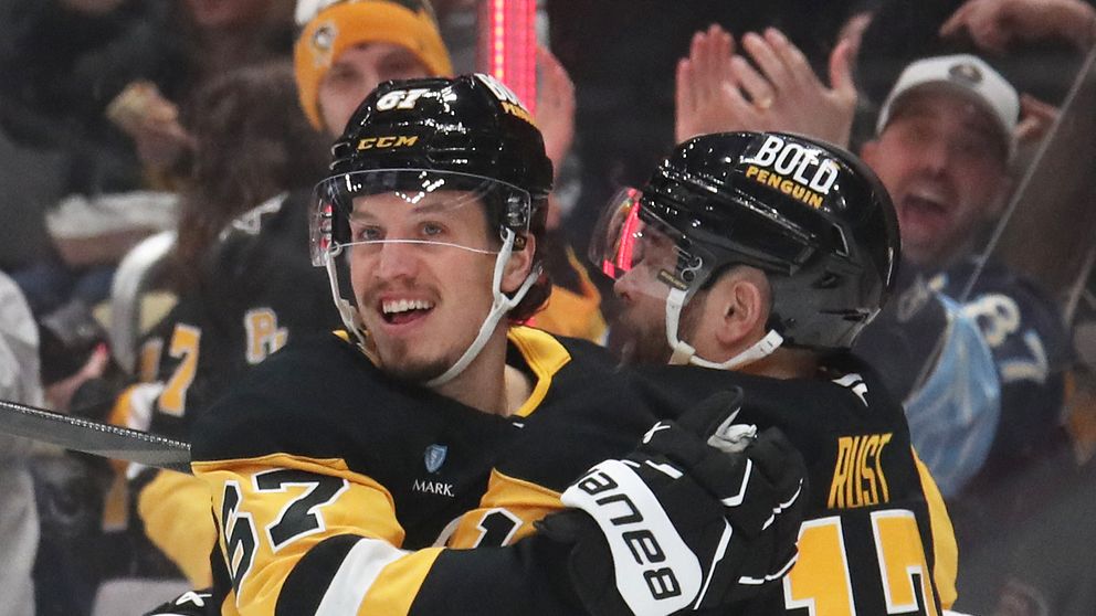 Spelare från Pittsburgh Penguins kramas och firar en målscore under en match, med jublande åskådare i bakgrunden. De bär sina svarta och gula matchtröjor, och stämningen är energisk och glad.