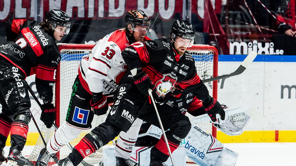 En intensiv hockeysituation på isen där en spelare i svart och röd utrustning försöker kontrollera pucken medan en motståndare i vit och grön utrustning trycker på. I bakgrunden syns en målvakt i position för att försvara målet.