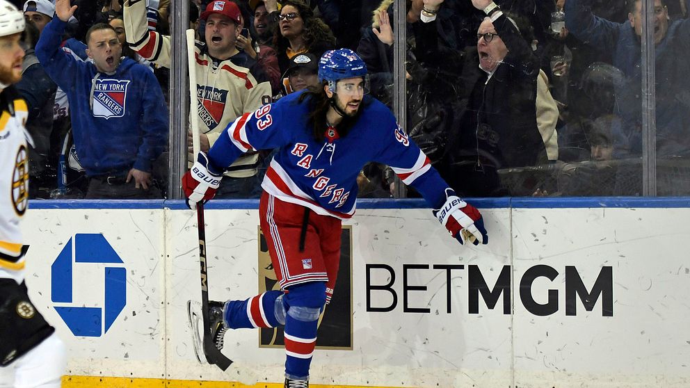 Hockeyspelaren i New York Rangers-tröja firar ett mål vid sargen, medan åskådarna hejar på i bakgrunden. I fokus finns även reklamskylten för BETMGM på glaset vid spelområdet.