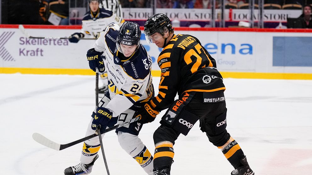Ishockeyspelare från två olika lag, det ena i mörka dräkter och det andra i ljusa, kämpar om pucken under en match.
