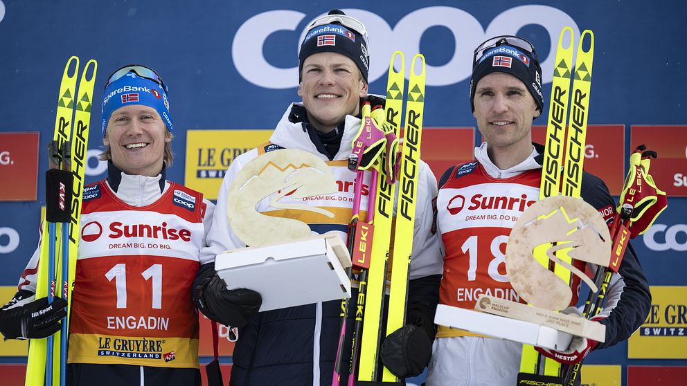 Tre skidåkare står på prispallen efter en tävling i Engadin med sina skidor och troféer. De är klädda i tävlingsdräkter och har nummerlappar med nummer 11 och 18. Skidorna har märket Fischer.