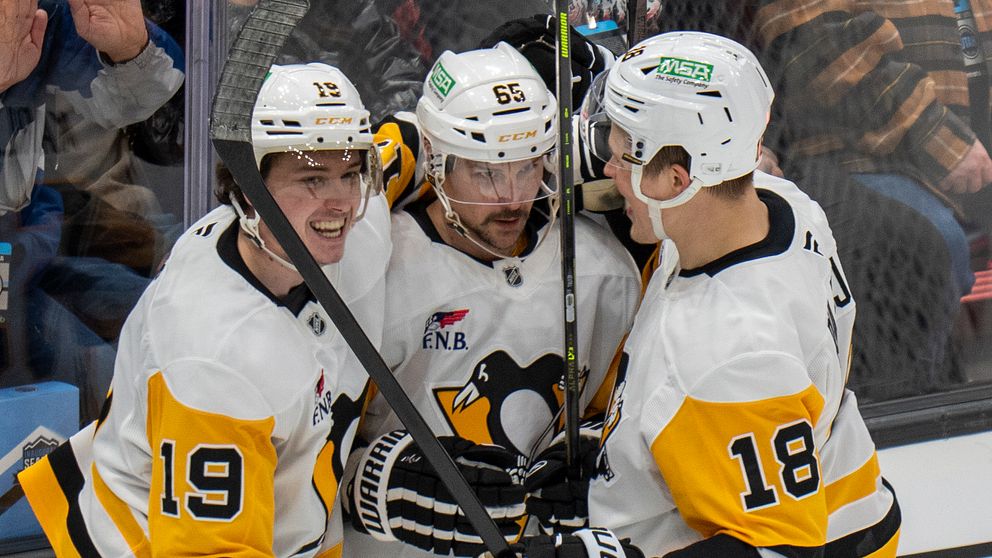 Tre ishockeyspelare i vit och gul mundering från Pittsburgh Penguins firar ett mål tillsammans på isen medan åskådare syns i bakgrunden.