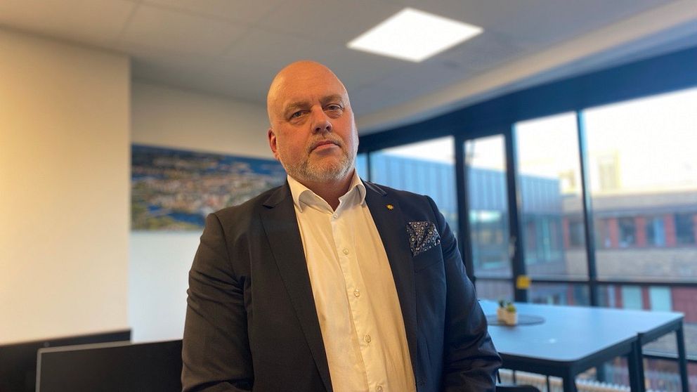 En person i kostym står inomhus i ett kontorsutrymme, med stora fönster i bakgrunden som ger utsikt över byggnader utanför.