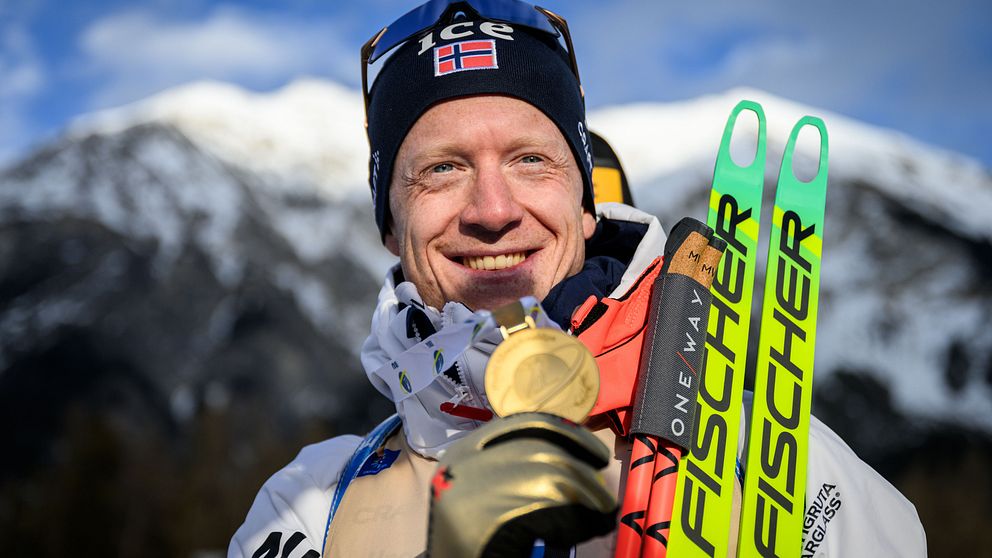 En skidåkare iförd hjälm och skidutrustning håller upp en guldmedalj framför ett snöklätt bergslandskap, med ett par Fischer-skidor i ena handen.