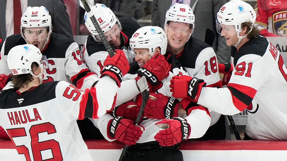Spelare från ett hockeylag firar ett mål från Jesper Bratt på bänken. De kramar och klappar om en lagkamrat, medan andra spelare ser glada ut och jublar. Lagets röda och vita dräkter dominerar bilden, vilket skapar en känsla av gemenskap och framgång.