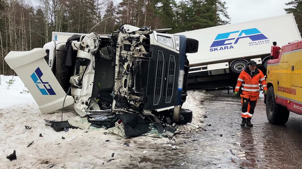 I bilden syns en lastbil som är krockad framtill som ligger på sidan i snön. Till höger går en man i orange klädsel. I bakgrunden syns en till lastbil som ligger i diket.