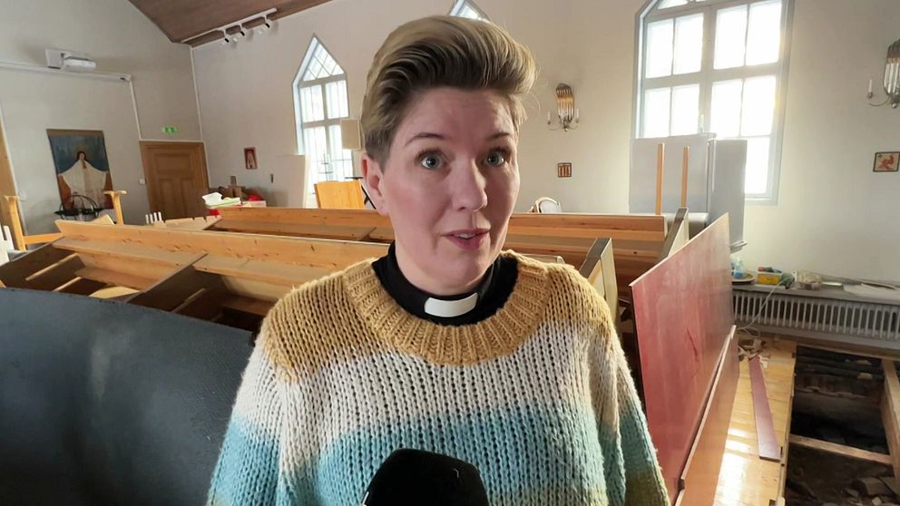 En person iklädd en stickad tröja och en prästkrage står i en kyrka med träbänkar och ljusa fönster i bakgrunden.