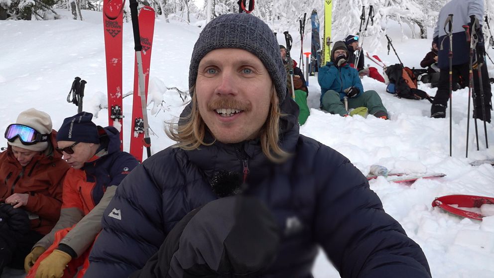 En grupp människor klädda i vinterkläder sitter och fika i en snöig miljö med skidor uppställda i bakgrunden. De är omgivna av snötäckta träd, vilket antyder att de befinner sig på eller nära en skidort.