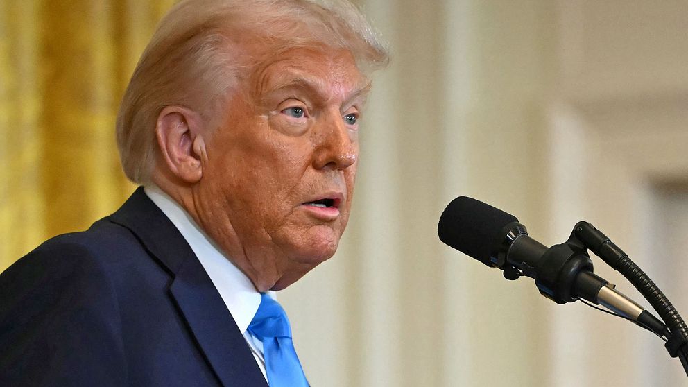 Donald Trump står vid en mikrofon i ett inomhusmiljö, med guldfärgad bakgrund.