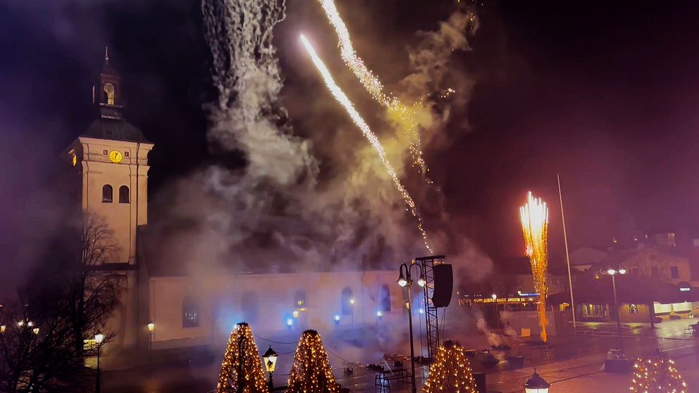 Eldverken lyser upp natthimlen över Varbergs stadskärna, med en klocktorn i bakgrunden. Rök och ljus skapar en festlig atmosfär, medan fyrverkerier exploderar och sprayar färg över scenen. En del av torgen är dekorerade med julbelysning, vilket ytterligare förstärker den festliga stämningen.