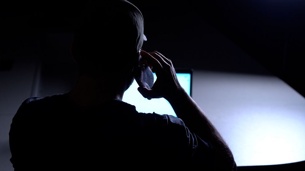 En man fotad bakifrån, sitter och pratar i mobiltelefon framför en datorskärm.