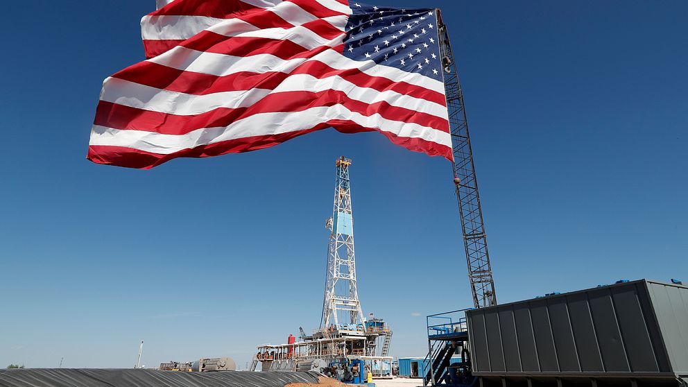 Amerikanska flaggan vajar i vinden framför en oljeplattform som syns i bakgrunden, med blå himmel som bakgrund.