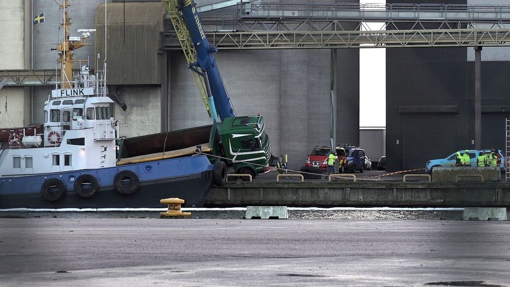 En båt ligger vid en kaj där en grön lastbil har stött mot båten. En kran arbetar för att lyfta lasten från lastbilen, medan flera personer i reflexvästar observerar och assisterar i operationen. I bakgrunden syns byggnader och en industriell miljö.