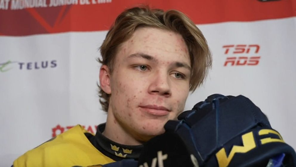 En ung ishockeyspelare iförd en gul tröja står framför en backdrop med sponsormärken efter en match. Han håller i sina handskar med fokus på den kommande intervjun.