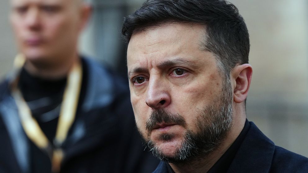 Volodymyr Zelenskyj i mörk jacka, med suddat ansikte, utomhus med en person i bakgrunden
