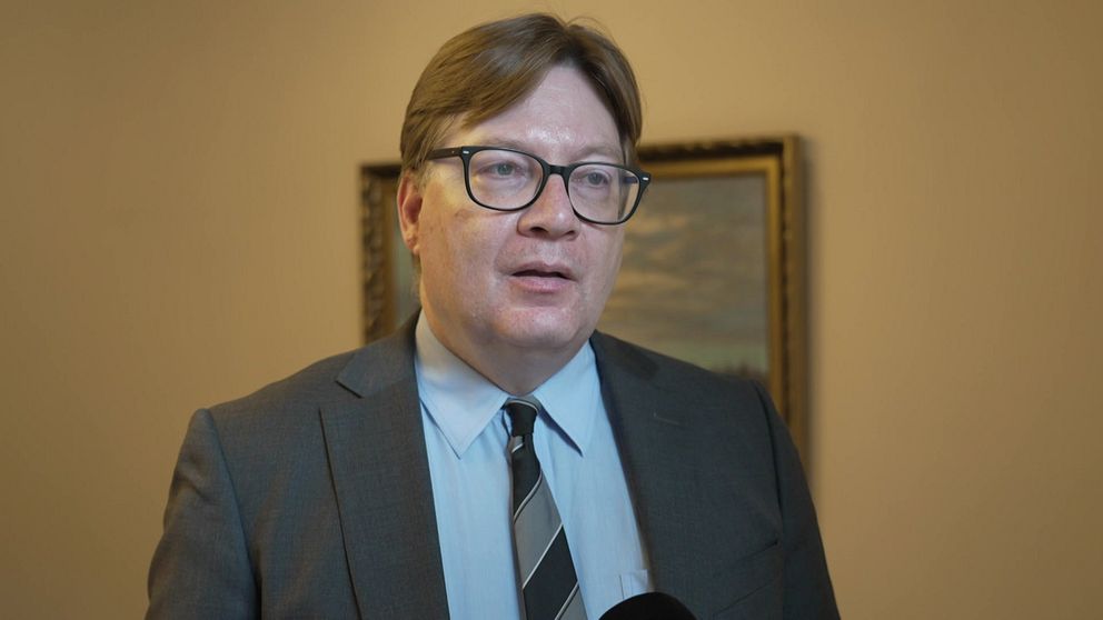 Man i kostym och slips intervjuas inomhus framför en tavla.