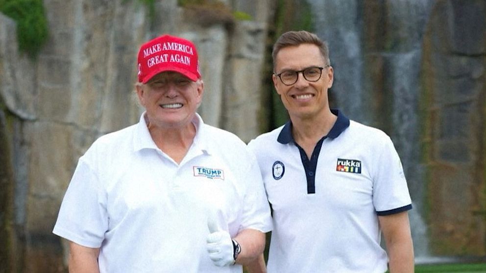Två personer i vita skjortor vid stenmur. En har röd keps med slogan ”Make America Great Again”.