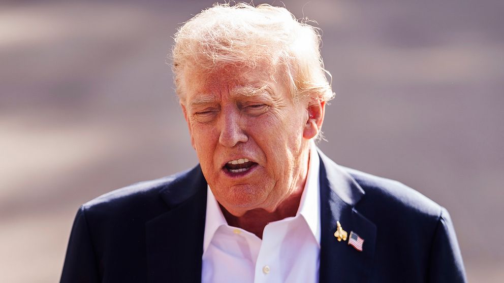 Stillbild: Donald Trump utomhus i kostym och vit skjorta, amerikansk flaggnål på kavajen.