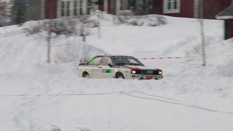 En rallybil kör genom ett snötäckt landskap i en vinterrallytävling. I bakgrunden syns ett par hus och träd täckta av snö.