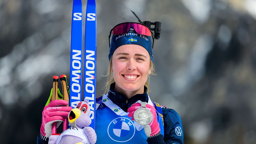 En idrottare klädd i tävlingsdräkt håller upp ett par blå Salomon-skidor och en medalj i ena handen och en lila gosedjursfågel i den andra. Skidgrenar är synliga i bakgrunden.