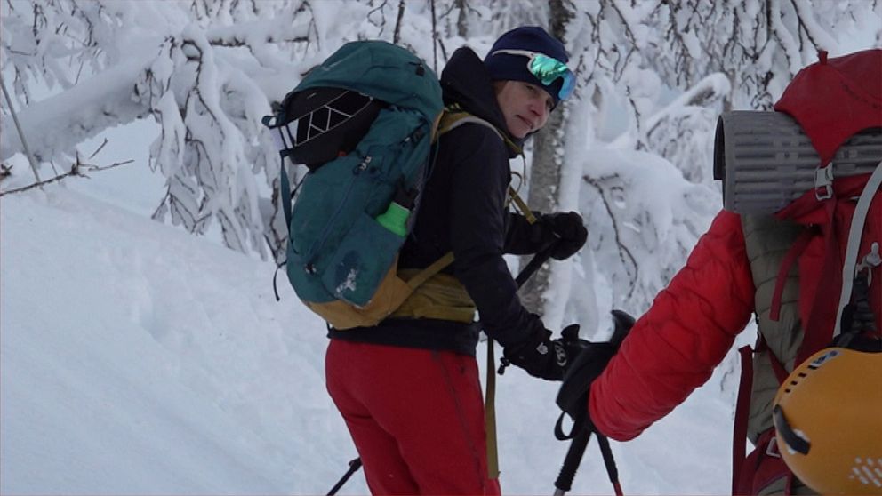 Två personer i vinterkläder och med ryggsäckar på ryggen åker skidor i ett snötäckt skogslandskap.