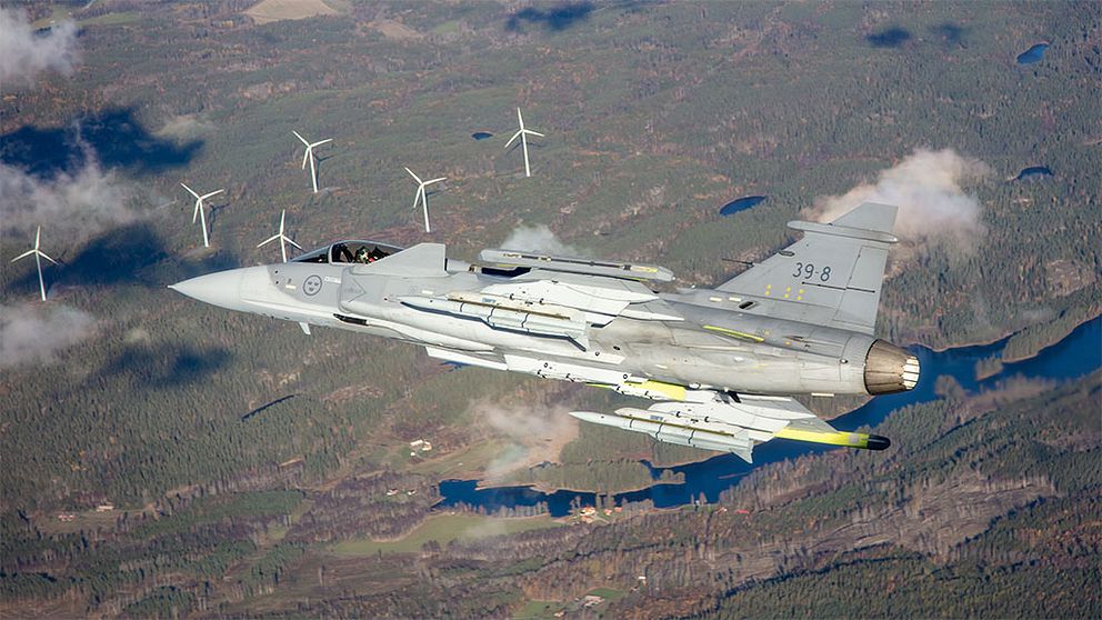 Stridsflygplanet Gripen E flyger över landskap med vindkraftverk i bakgrunden.