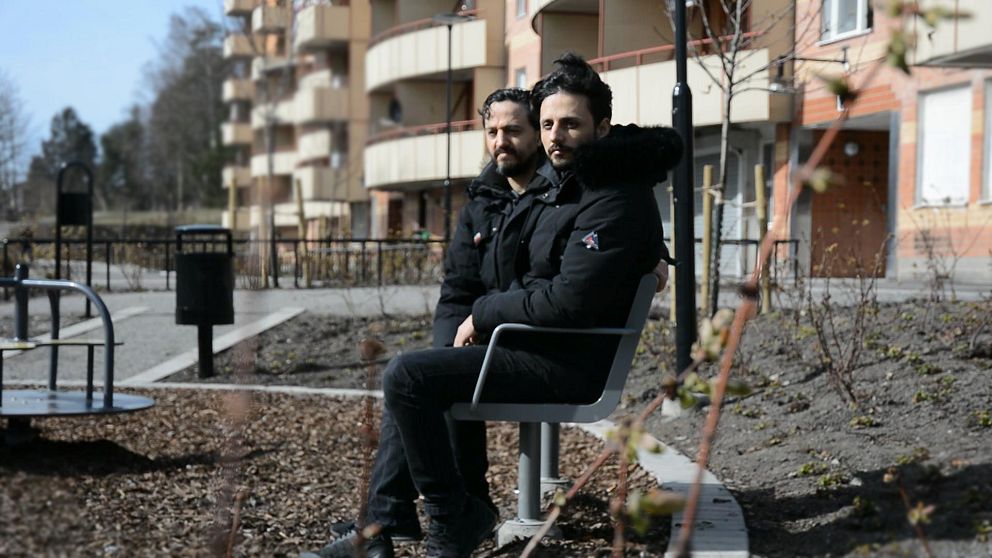Två personer sitter på en bänk i en urban miljö, omgivna av flerfamiljshus och en välordnad parklandskap. Det är en solig dag med snålt med vegetation i bakgrunden.