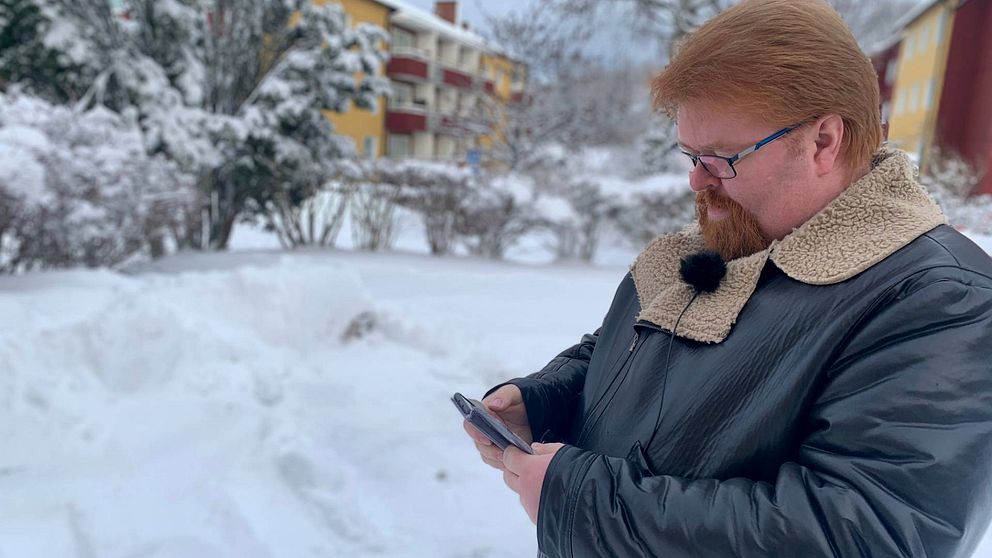 Person i vinterkläder använder telefon utomhus, snötäckta buskar i bakgrunden.