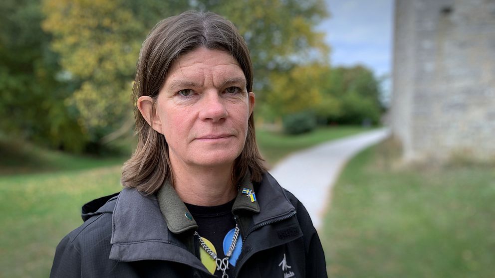 Person med långt hår står utomhus. Bakom syns en grönskande park och en stenbyggnad.