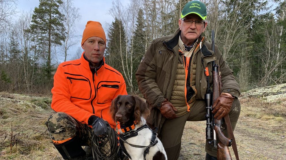 Två jägare i skogen med en hund mellan sig.
