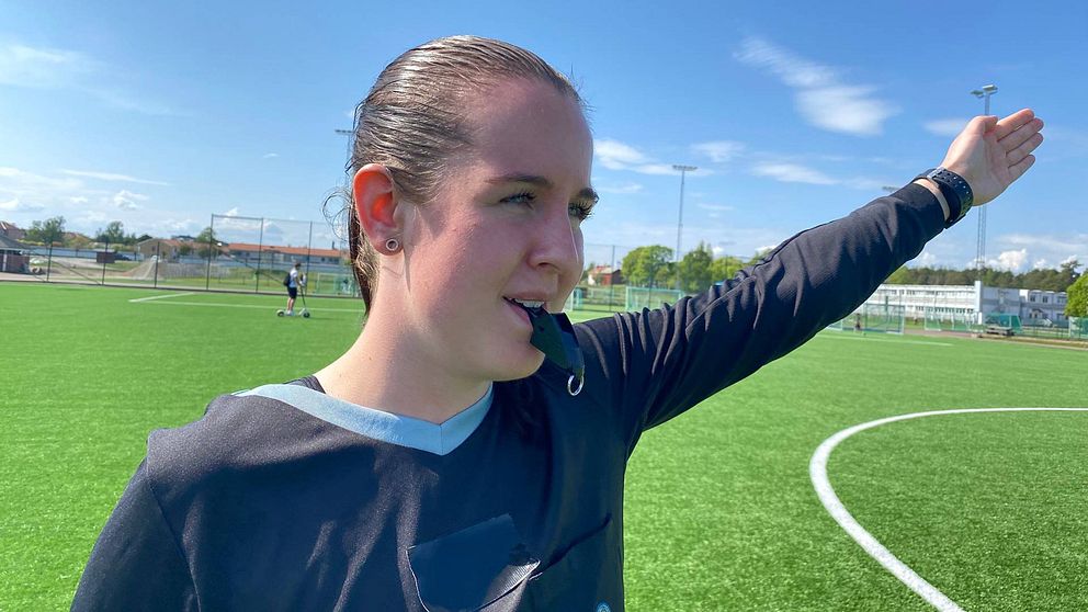 En kvinnlig fotbollsdomare som är svartklädd och håller pipan i munner samtidigt som hon pekar med sin vänstra arm.