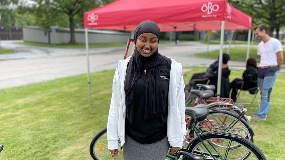 En ung tjej står och ler och tittar rakt in i kameran. I bakgrunden står några cyklar samtidigt som några personer sitter och pratar och skrattar.