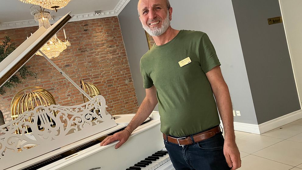 En man i grön t-tröja står vid sidan av ett stort vit piano med sin högra hand på det och tittar in mot kameran med ett leende.