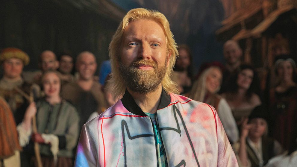 Ljusblond man med skägg ler in i kameran. Suddig bakgrund med människor.